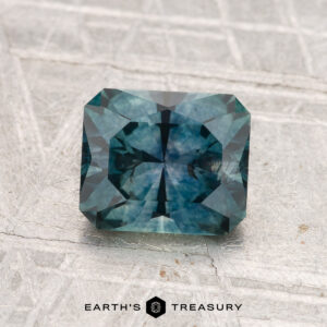 A 1.59-carat heated Montana sapphire with a rectangular step cut and blue-green hue rests on a textured gray surface, with "EARTH'S TREASURY" below it.