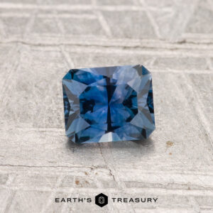 A rectangular, faceted blue 0.80-carat Montana sapphire (heated) is displayed on a textured, light surface with "EARTH'S TREASURY" at the bottom.