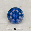A 1.72-carat heated Montana sapphire, round brilliant-cut and blue, is displayed on a light textured surface with "EARTH'S TREASURY" below.