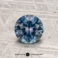 A 1.32-carat heated Montana sapphire with a round brilliant cut and blue-green hue rests on a textured gray surface, with "EARTH'S TREASURY" at the bottom.