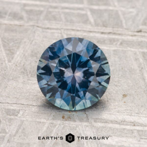 A 1.32-carat heated Montana sapphire with a round brilliant cut and blue-green hue rests on a textured gray surface, with "EARTH'S TREASURY" at the bottom.