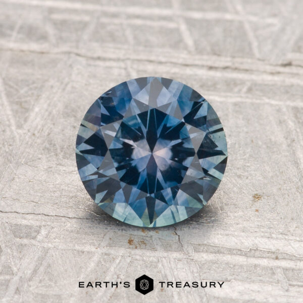 1.32-Carat Montana Sapphire (Heated) A 1.32-carat heated Montana sapphire with a round brilliant cut and blue-green hue rests on a textured gray surface, with "EARTH'S TREASURY" at the bottom.