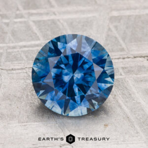 A 2.19-carat heated Montana sapphire, round brilliant cut, is displayed on a textured gray surface with "EARTH'S TREASURY" text at the bottom.