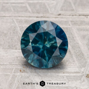 1.78-Carat Montana Sapphire (Heated), round brilliant-cut, displayed on a textured gray surface with the "Earth's Treasury" logo and text beneath the stone.