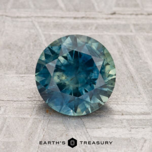 A 1.71-carat heated Montana sapphire, round and faceted in blue-green, is displayed on a textured gray surface with "Earth's Treasury" at the bottom.