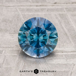 A 1.70-carat round brilliant-cut heated Montana sapphire is displayed upright on a textured gray surface, with the "Earth's Treasury" logo shown at the bottom.