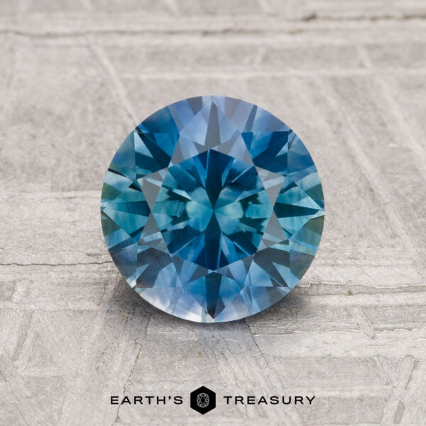 1.70-Carat Montana Sapphire (Heated) A 1.70-carat round brilliant-cut heated Montana sapphire is displayed upright on a textured gray surface, with the "Earth's Treasury" logo shown at the bottom.