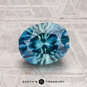 A 2.05-carat heated Montana sapphire is displayed on a textured gray surface, with the "Earth's Treasury" logo at the bottom.