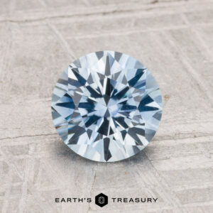 A 1.64-carat heated Montana sapphire, round brilliant-cut and blue in color, is shown on a gray textured surface with "EARTH'S TREASURY" written below.