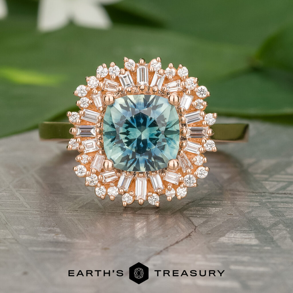 The _SNY1574-Edit-1200-BW gold ring features a round blue-green gemstone at the center, accented by a halo of baguette and round diamonds, shown on a patterned surface.