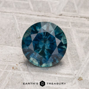 A round, deep blue-green 1.42-carat Montana Sapphire (Heated) is displayed on a textured light surface with "EARTH'S TREASURY" below.