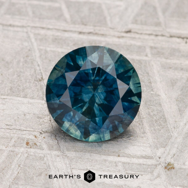 1.42-Carat Montana Sapphire (Heated) A round, deep blue-green 1.42-carat Montana Sapphire (Heated) is displayed on a textured light surface with "EARTH'S TREASURY" below.