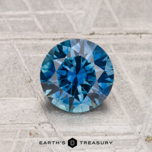 1.30-carat heated Montana sapphire, round brilliant-cut, showcased on a textured gray surface with "EARTH'S TREASURY" text at the bottom.