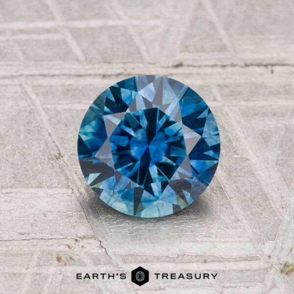 1.30-Carat Montana Sapphire (Heated) 1.30-carat heated Montana sapphire, round brilliant-cut, showcased on a textured gray surface with "EARTH'S TREASURY" text at the bottom.