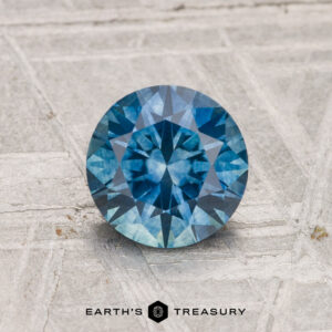 A 1.29-carat heated Montana Sapphire, round and faceted, rests on a textured gray surface with "Earth's Treasury" below it.