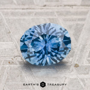 A 1.91-carat oval-cut Montana sapphire (heated) is displayed on a textured gray surface with the text "EARTH'S TREASURY" beneath it.