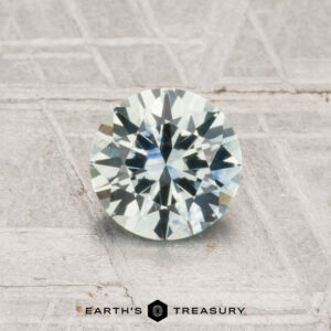 A 1.03-carat, round brilliant-cut Montana sapphire (heated) is displayed on a textured gray surface with “Earth’s Treasury” at the bottom.