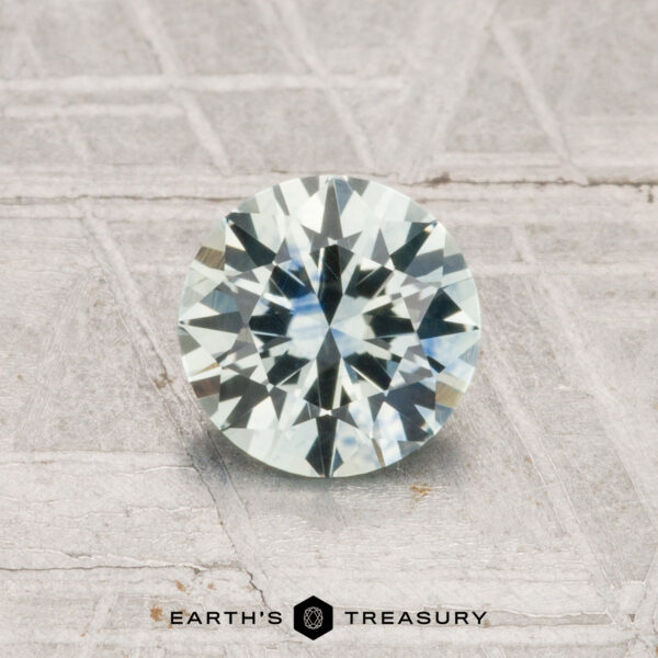 1.03-Carat Montana Sapphire (Heated) A 1.03-carat, round brilliant-cut Montana sapphire (heated) is displayed on a textured gray surface with “Earth’s Treasury” at the bottom.