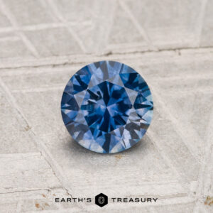 A round brilliant-cut 0.79-carat heated Montana sapphire is shown on a light textured surface with "Earth's Treasury" at the bottom.