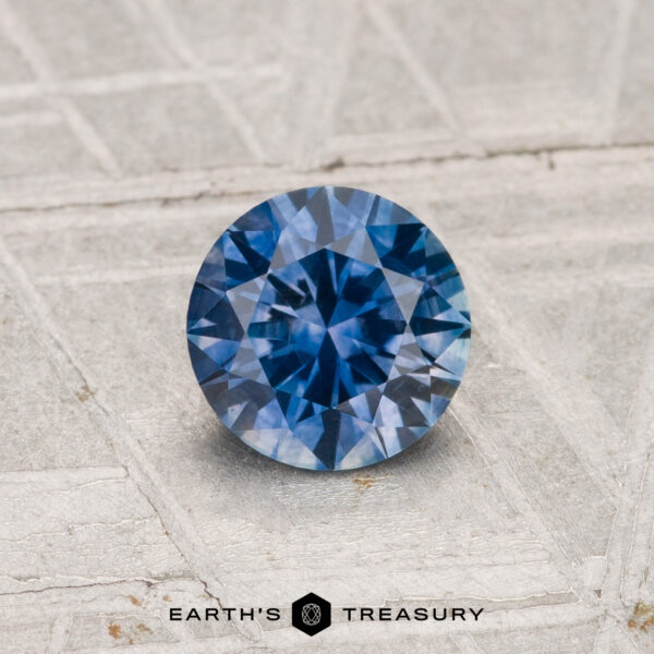 0.79-Carat Montana Sapphire (Heated) A round brilliant-cut 0.79-carat heated Montana sapphire is shown on a light textured surface with "Earth's Treasury" at the bottom.