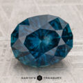 A 4.96-carat heated Montana sapphire, cut in a faceted oval shape and vibrant blue, is showcased on a textured surface with the "Earth's Treasury" text and logo beneath it.