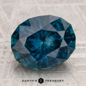 A 4.96-carat heated Montana sapphire, cut in a faceted oval shape and vibrant blue, is showcased on a textured surface with the "Earth's Treasury" text and logo beneath it.