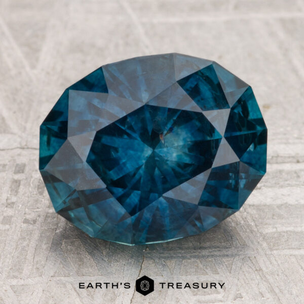 A 4.96-carat heated Montana sapphire, cut in a faceted oval shape and vibrant blue, is showcased on a textured surface with the "Earth's Treasury" text and logo beneath it.