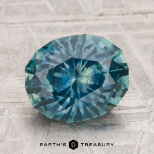 A 3.50-carat heated Montana sapphire, cut in a faceted oval shape with blue-green hues, displayed on a textured surface. “EARTH'S TREASURY” appears at the bottom.