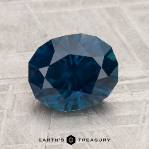 A 2.50-carat heated Montana sapphire, faceted and oval-shaped, is displayed on a textured gray surface with "EARTH'S TREASURY" at the bottom.