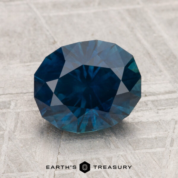 2.50-Carat Montana Sapphire (Heated) A 2.50-carat heated Montana sapphire, faceted and oval-shaped, is displayed on a textured gray surface with "EARTH'S TREASURY" at the bottom.