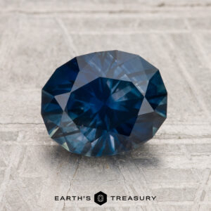 A 2.38-carat heated Montana Sapphire with a round brilliant cut and deep blue color is displayed on a textured gray surface, with "EARTH'S TREASURY" shown at the bottom.
