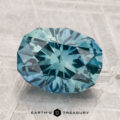 A 3.93-carat heated Montana sapphire, faceted in an oval shape with blue-green hues, rests on a textured gray surface above the "EARTH'S TREASURY" text and hexagon logo.