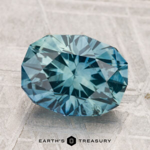 A 3.93-carat heated Montana sapphire, faceted in an oval shape with blue-green hues, rests on a textured gray surface above the "EARTH'S TREASURY" text and hexagon logo.