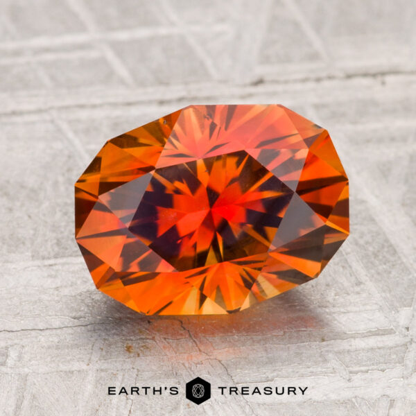 A 3.09-carat heated Montana sapphire with orange hues and red-yellow flashes is showcased on a textured grey surface, with "EARTH'S TREASURY" displayed below.