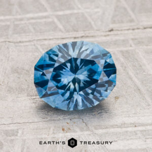 A 1.68-carat heated Montana sapphire, cut in a faceted oval shape, is displayed on a textured gray surface with "EARTH'S TREASURY" at the bottom.