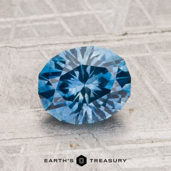 1.68-Carat Montana Sapphire (Heated) A 1.68-carat heated Montana sapphire, cut in a faceted oval shape, is displayed on a textured gray surface with "EARTH'S TREASURY" at the bottom.