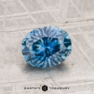 A faceted oval 1.60-carat heated Montana sapphire is displayed on a textured gray surface, with the text "EARTH'S TREASURY" at the bottom.
