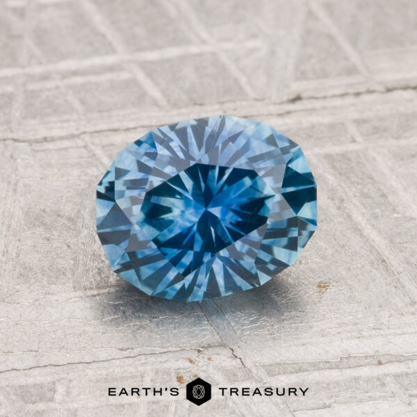 1.60-Carat Montana Sapphire (Heated) A faceted oval 1.60-carat heated Montana sapphire is displayed on a textured gray surface, with the text "EARTH'S TREASURY" at the bottom.
