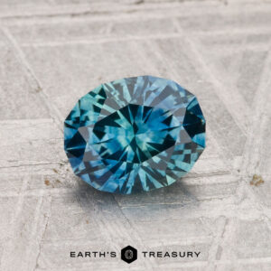 A 1.34-carat heated Montana sapphire, faceted in an oval shape with a blue-green hue, rests on a textured gray surface above the text "EARTH'S TREASURY.