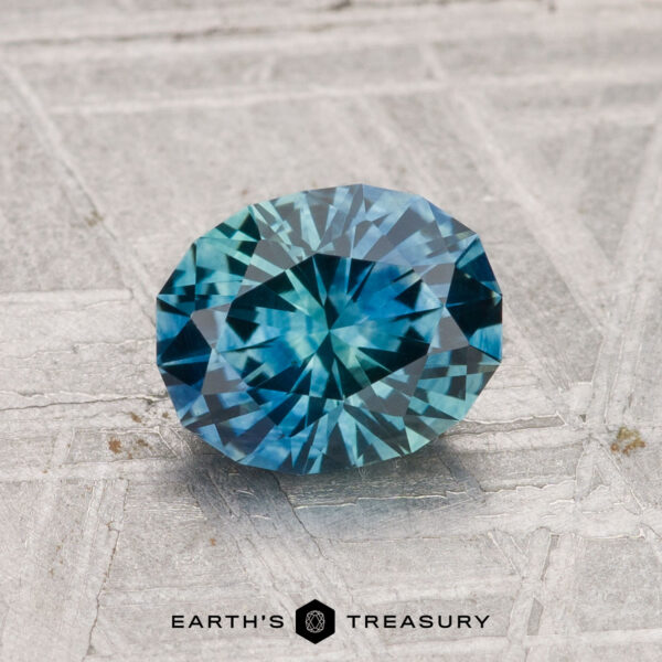 1.34-Carat Montana Sapphire (Heated) A 1.34-carat heated Montana sapphire, faceted in an oval shape with a blue-green hue, rests on a textured gray surface above the text "EARTH'S TREASURY.
