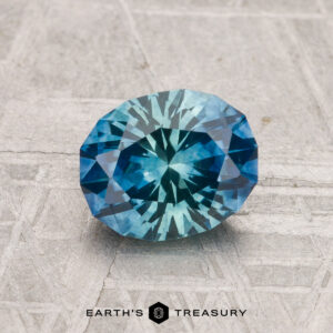 A 1.50-carat heated Montana sapphire, cut in a faceted oval shape, rests on a textured gray surface with "EARTH'S TREASURY" displayed at the bottom.
