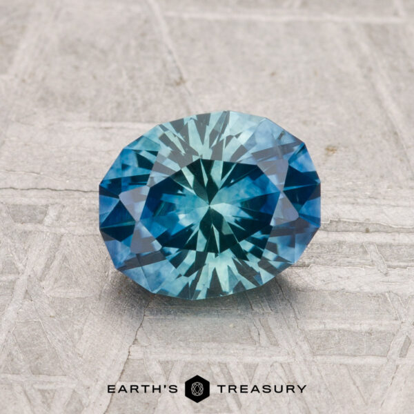 1.50-Carat Montana Sapphire (Heated) A 1.50-carat heated Montana sapphire, cut in a faceted oval shape, rests on a textured gray surface with "EARTH'S TREASURY" displayed at the bottom.