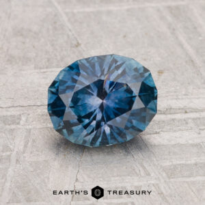 A 1.33-carat heated Montana sapphire, round brilliant-cut and blue, is displayed on a textured gray surface with the "Earth's Treasury" logo at the bottom.