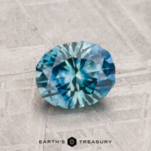 A 1.39-carat heated Montana sapphire, round and faceted with a blue-green hue, is displayed on a textured gray surface featuring "EARTH'S TREASURY" and a logo at the bottom.