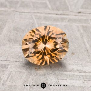 A 1.37-carat, round, faceted golden-brown Montana sapphire (heated) is displayed on a textured gray surface with "EARTH'S TREASURY" at the bottom.