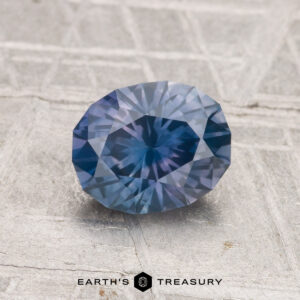 A 1.37-carat heated Montana sapphire, faceted in an oval shape and blue in color, is displayed on a textured gray surface with "EARTH'S TREASURY" at the bottom.