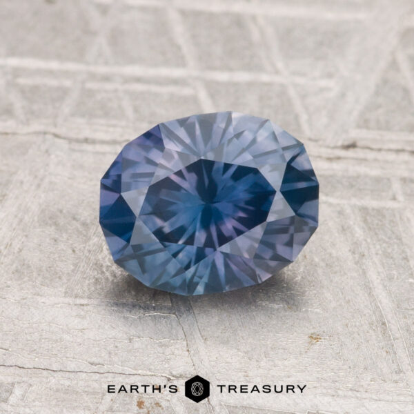 1.37-Carat Montana Sapphire (Heated) A 1.37-carat heated Montana sapphire, faceted in an oval shape and blue in color, is displayed on a textured gray surface with "EARTH'S TREASURY" at the bottom.