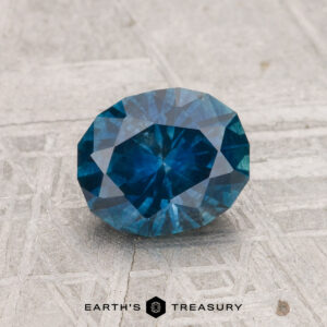 A 1.36-carat heated Montana sapphire with an oval cut is displayed on a textured gray surface, with "EARTH'S TREASURY" visible at the bottom.