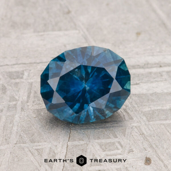 1.36-Carat Montana Sapphire (Heated) A 1.36-carat heated Montana sapphire with an oval cut is displayed on a textured gray surface, with "EARTH'S TREASURY" visible at the bottom.