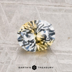 A 1.29-carat heated Montana sapphire, round and faceted with yellow and clear color zoning, rests on a textured gray surface. "Earth's Treasury" is shown at the bottom.